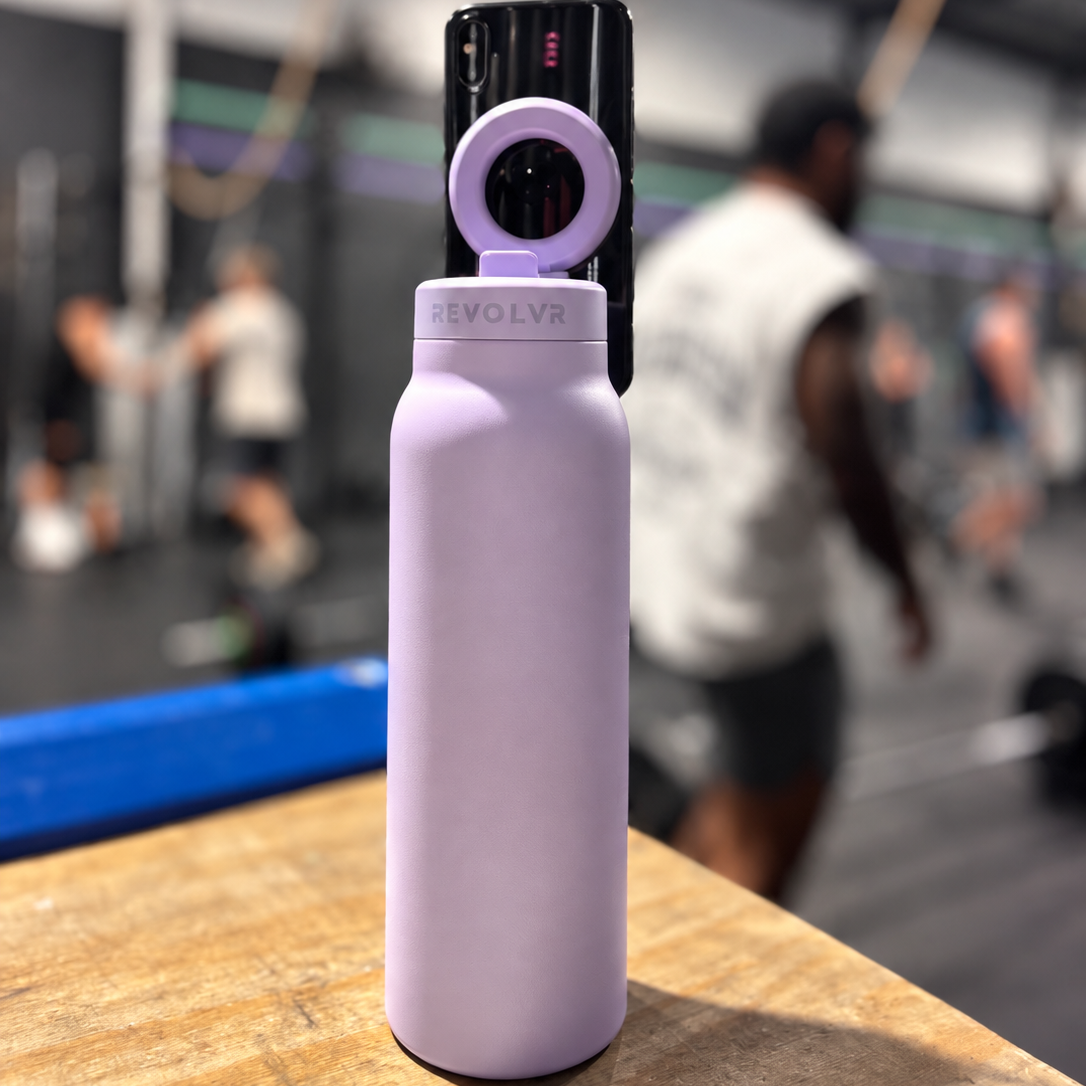 A light purple insulated water bottle with a black and purple phone mount attached, standing on a wooden surface in a blurred gym setting.
