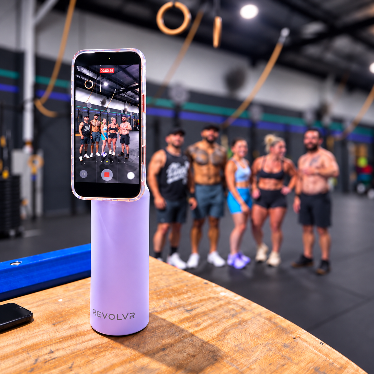 A light purple REVOLVR phone stand holding a smartphone in a gym setting, with a group of people standing blurred in the background.