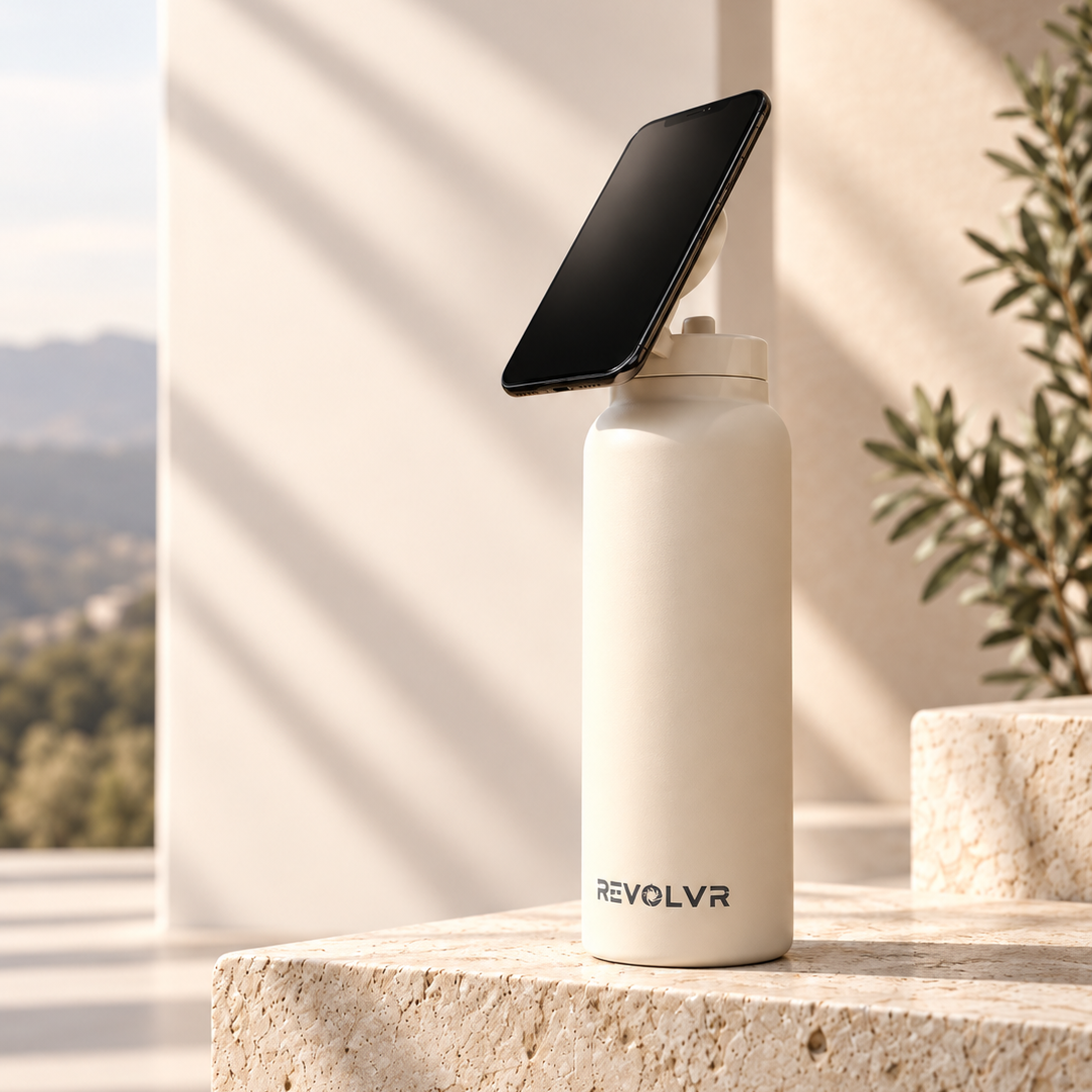 A REVOLVR insulated bottle in warm off-white stands on a stone ledge outdoors, with a black smartphone propped against the bottle on a matching holder. Sunlight and shadows fall across a beige architectural background with a blurred plant and distant landscape.
